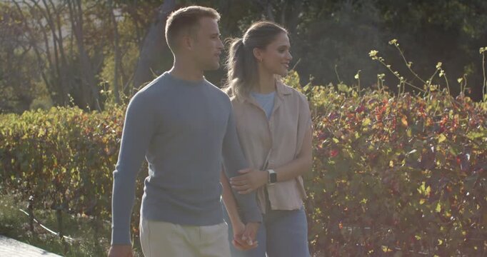 Diverse couple walking arm-in-arm along hedge path, man pointing at hill, woman wearing smartwatch