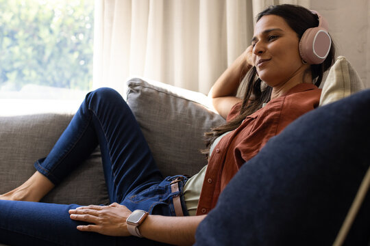 Indian woman lounging on gray couch at home, wearing pink headset, rust shirt, jeans, watch
