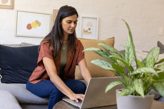 Indian woman sitting on gray sofa typing on laptop near potted variegated plant, copy space