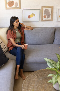 Indian woman sitting barefoot on sofa in green top holding pink mug, phone, talking, copy space