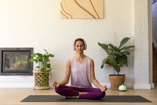 Adult woman meditating on yoga mat on wood floor, wearing burgundy leggings and headphones