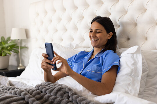 Indian woman wearing blue pajama set with white piping reclining on bed tapping smartphone