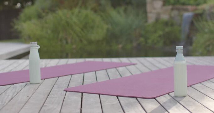 Pink mats and white bottles are resting on deck while breeze is rippling pond, awaiting yoga