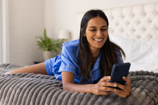 Indian woman lying on stomach on bed wearing blue satin pajamas, using dark smartphone and smiling