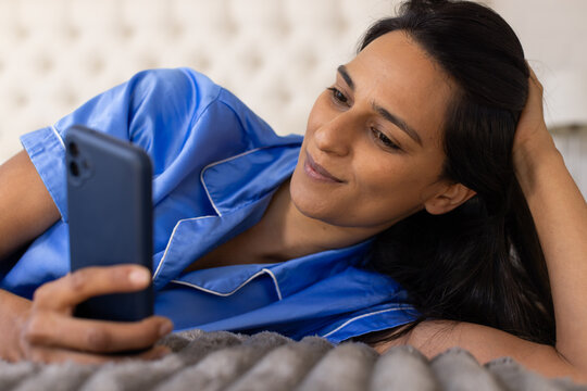 Indian woman reclining on bed, holding smartphone, propping head on hand, wearing blue satin top