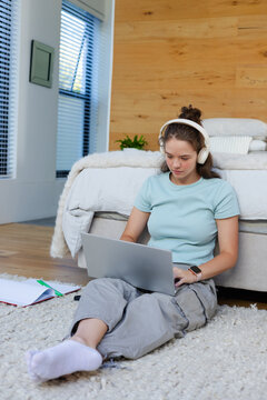 Female adult sitting on cream shag rug in bedroom, studying on silver laptop with headphones