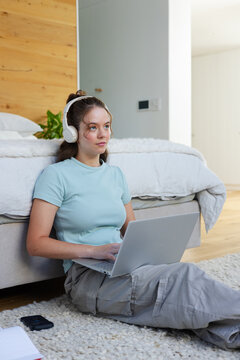 Woman sitting on shaggy bedroom rug, wearing headphones and light-blue top, typing on laptop