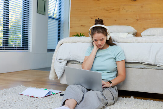 Woman sitting on shag rug at foot of bed in bedroom, wearing headphones, using silver laptop