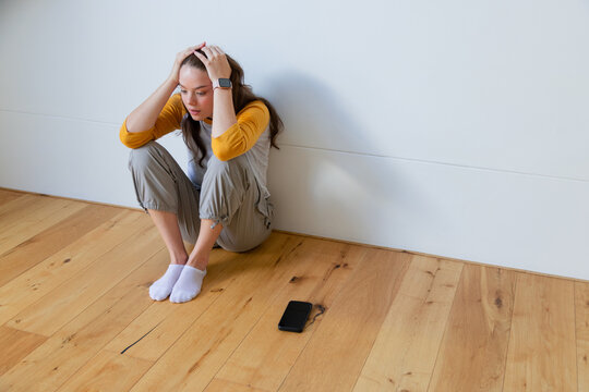 Adult woman sitting holding head on floor at home wearing yellow top, phone, smartwatch, copy space