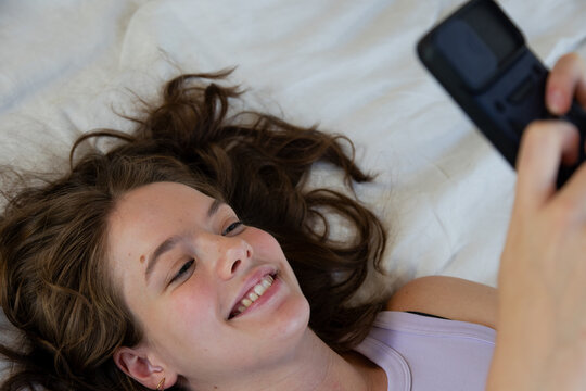 Female lying on white bedding smiling while holding clamshell phone and wearing light purple top