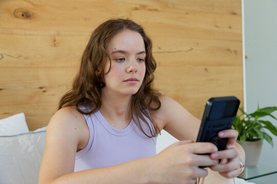 Female sitting on bed in bedroom, holding handheld device, wearing light purple sleeveless top