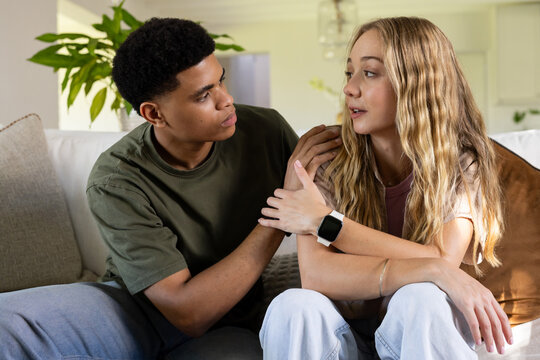 Diverse couple sitting on light sofa in living room, man placing hand on woman wearing smartwatch