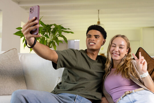 Diverse couple sitting on light sofa in bright living room taking pink smartphone selfie