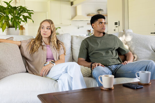 Diverse couple sitting apart on sofa in open-plan living room, man in olive-green tee, ceramic mugs