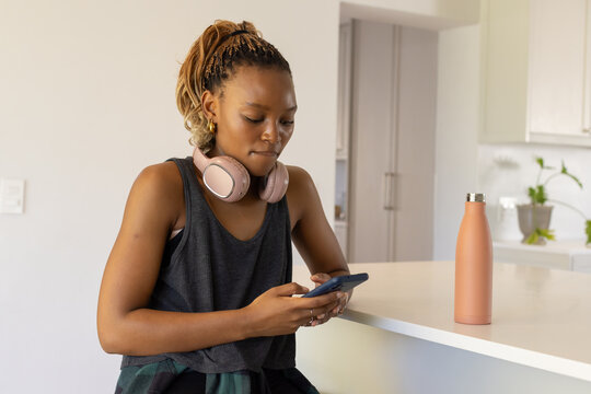 African American woman leaning against kitchen counter, using smartphone with pink headphones