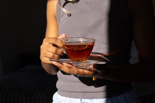 African woman cradling amber tea in glass teacup on saucer in sunlit bedroom in ribbed tanktop