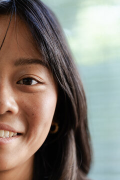 Asian female smiling showing skin texture, teeth, gold hoop earring and dark hair near blinds
