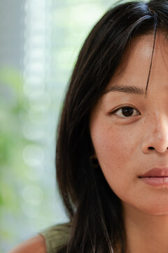 Asian woman posing calmly in room by blurred blinds with hoop earring, freckles, olive green strap