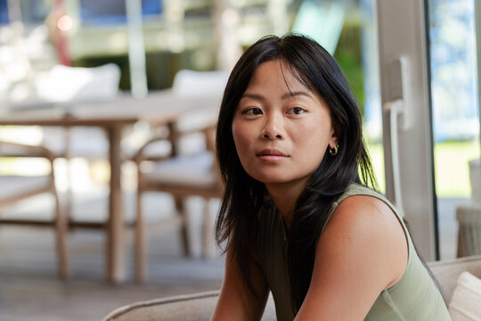 Asian woman sitting on sofa in modern lounge by glass sliding doors wearing olive top