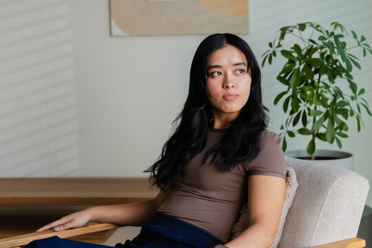 African American woman sitting on upholstered armchair in living room, wearing fitted brown top