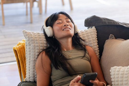 Adult Asian woman reclining on dark gray sofa at home, wearing white headphones holding smartphone