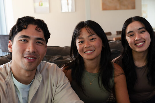 Three friends sitting close on sofa with cushions at home, wearing casual tops, pendant visible