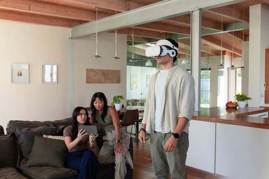Diverse friends watching man wearing white VR headset near kitchen, women holding tablet on sofa