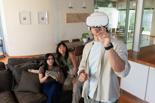 Diverse friends watching man wearing white VR headset gesturing at home with tablet, copy space