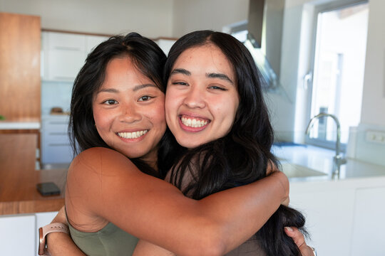 Asian and African American women hugging, smiling at camera in modern kitchen by counter with phone