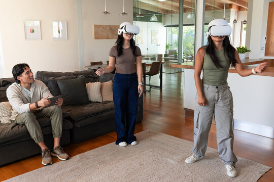 Diverse friends wearing white VR headsets and holding controllers in modern living room with tablet