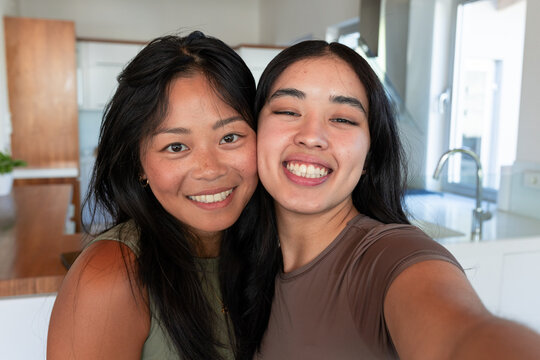 Diverse female friends leaning and smiling while holding phone and taking selfie in bright kitchen