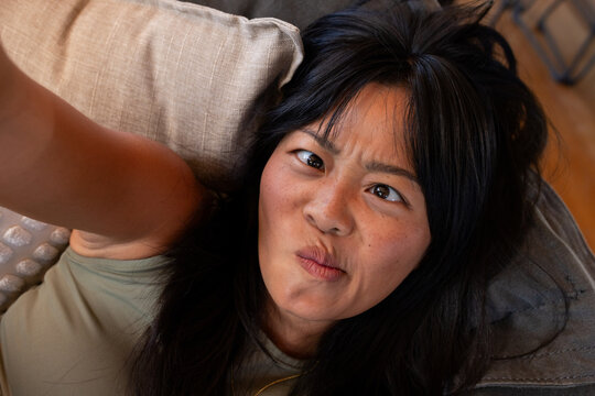 Asian woman in her twenties reclining on beige couch at home wearing olive-green top, holding phone
