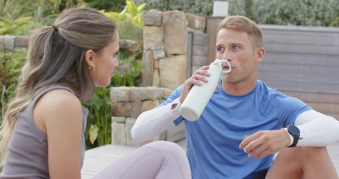 Diverse couple sitting on wooden deck man opening bottle cap and drinking, hydrating post workout