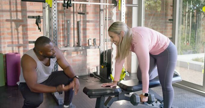 African American coach cueing as female client doing single-arm dumbbell rows on bench in home gym
