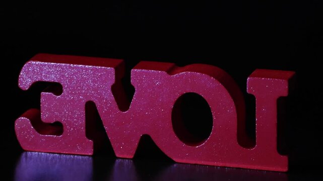 Red wooden "LOVE" text emerges from darkness, rotates from reversed to front view, then spins and fades into a soft bokeh blur on a black background. 4K 50fps