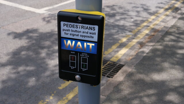 Close up of a British pedestrian crossing push button with illuminated blue WAIT signal and instruction panel on a city street pole, urban traffic safety infrastructure for people in the city.