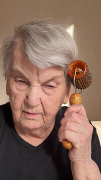Close-up senior woman massaging scalp with magnetic needle roller, focused expression, wooden handle contact, soft sunlight