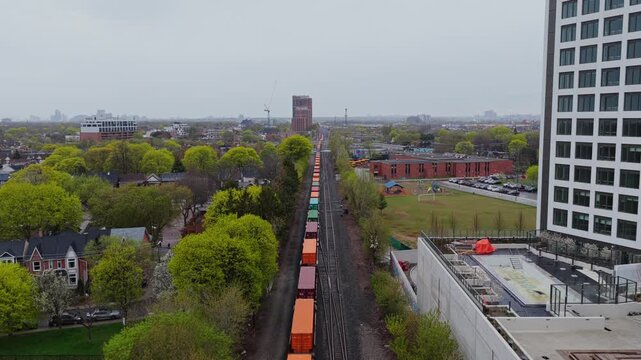 Freight train with intermodal containers moving on railway tracks through urban area and passing residential building with green park on overcast spring day in North America