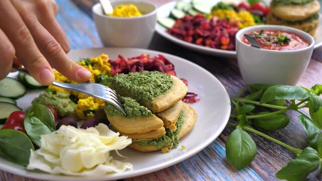 Human hands using a knife and fork to cut into savory vegan pancakes with green pesto, served with a healthy colorful salad and dips on a rustic blue wooden table in a restaurant setting