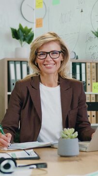 Successful beautiful business woman with glasses using tablet computer and working documents sitting at workplace in office looking at camera. Slow morion