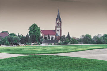 Neogotycki kościół z wysoką wieżą w otoczeniu zieleni. Panorama polskiej wsi, architektura sakralna i zielone pola. Spokojny krajobraz wiejski, pochmurne niebo, sielski klimat i natura. © Henryk Niestrój