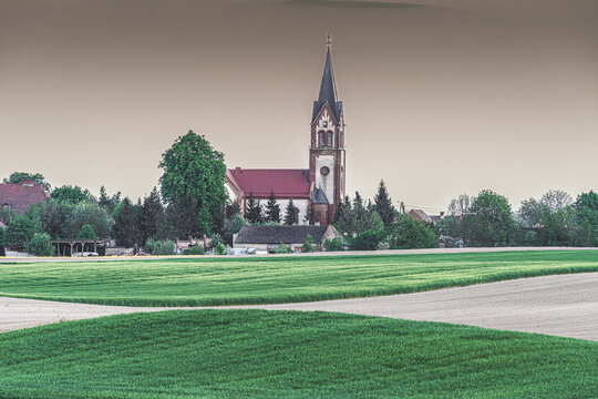 Neogotycki kości&oacute;ł z wysoką wieżą w otoczeniu zieleni. Panorama polskiej wsi, architektura sakralna i zielone pola. Spokojny krajobraz wiejski, pochmurne niebo, sielski klimat i natura.