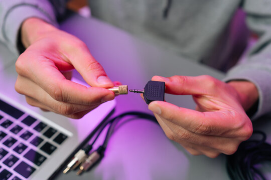 Hands connect audio jack adapter. Sound engineer or DJ sets up equipment with laptop. Modern workspace with cables under purple light. Technology, connectivity, music production concept.