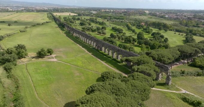 Aerial panoramic view of the Parco degli Acquedotti in Rome, Italy, featuring the long, ancient stone arches of a Roman aqueduct stretching across vibrant green fields toward the distant city skyline.