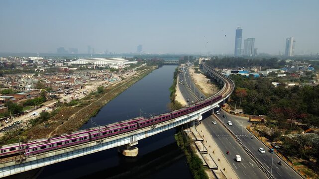 Aerial drone view of two metro trains crossing each other on elevated tracks alongside a river in Delhi city.