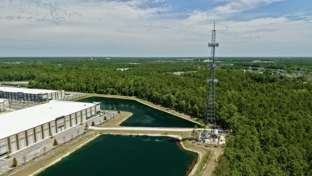 Aerial view of a tall telecommunications tower standing beside industrial warehouses, artificial ponds, and a dense green forest in World Golf Village, Florida, United States.