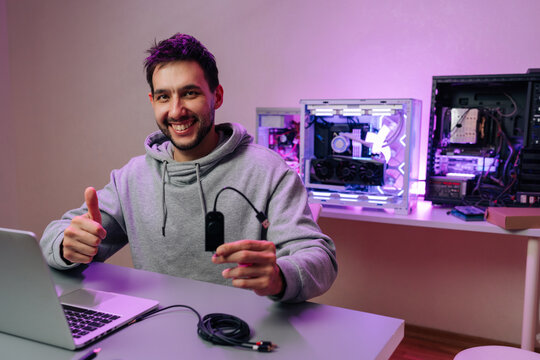 Happy technician holding hardware and giving thumbs up gesture. Man at desk with laptop and custom built computers with purple lighting. Concept for IT support, PC building, gaming, streaming.