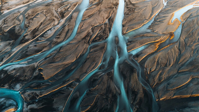 Aerial view of intricate braided glacial river patterns flowing through dark volcanic sand with golden sunlight highlights Skaftarhreppur, Iceland.