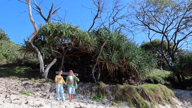 Young female friends enjoying a sunny day, standing on a sandy beach in australia and pointing at something, surrounded by lush pandanus trees and coastal vegetation under a clear blue sky