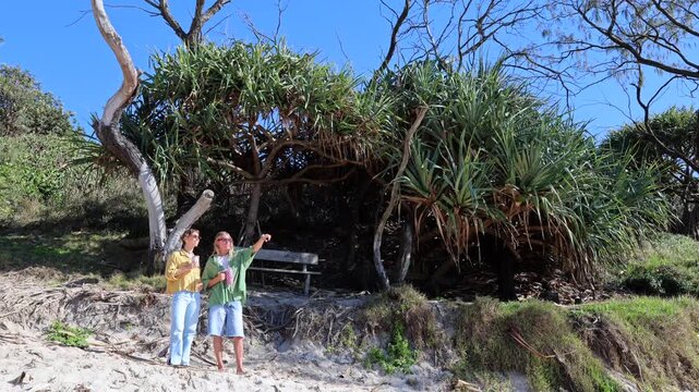 Young man and woman standing on a sandy dune, surrounded by lush pandanus trees. The man points towards something interesting while they both enjoy a beautiful sunny day in a natural setting